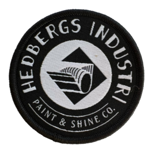 Patch med vår snygga logo Hedbergs Industri AB – Paint & Shine Co. Vävt tygmärke i svart med silvertråd. Framsida.